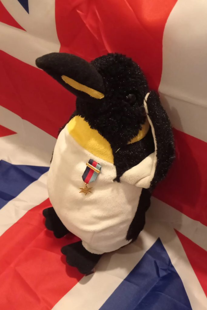 Photo d'une peluche manchot au garde à vous devant un drapeau du Royaume Uni. Il porte une médaille militaire sur le cœur. C'est Pinpin.