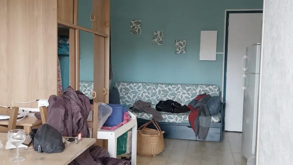 pièce avec canapé, frigo, armoire et table. Dessus, des vêtements, de la vaisselle, du bazar laissé en vrac. Au fond, une porte à moitié cachée.