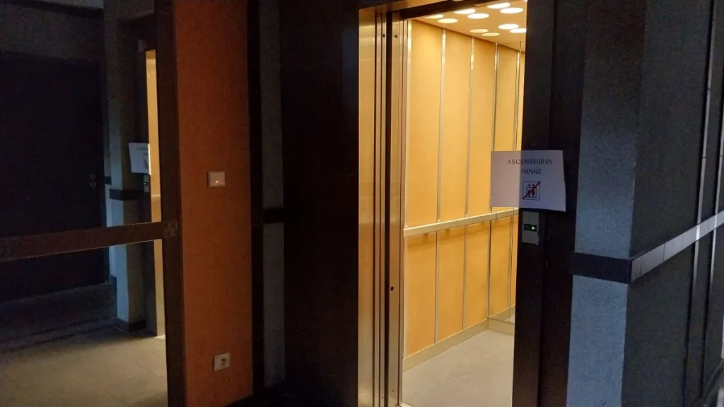 ascenseur ouvert dans un couloir plongé dans l'obscurité, avec un papier indiquant "ascenseur en panne"