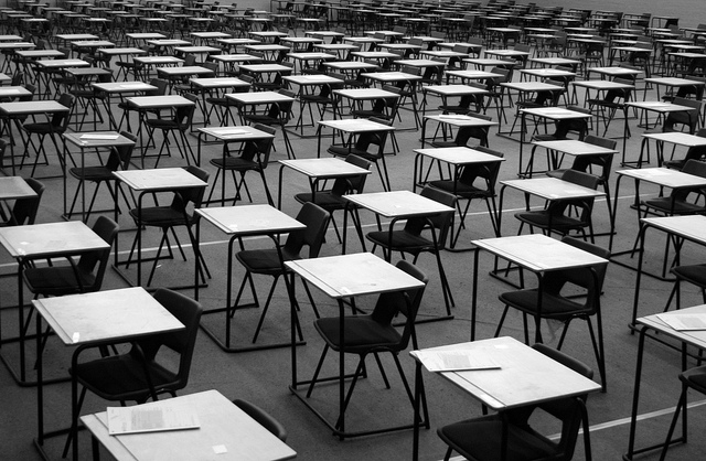 photo en noir et blanc de rangées de bureaux (et leurs chaises associées) à la manière d'une salle d'examen standard moderner (on voit ce qui ressemble à des sujets de composition sur les tables au premier plan). On ne voit pas de personnes ou êtres vivants dans la salle.